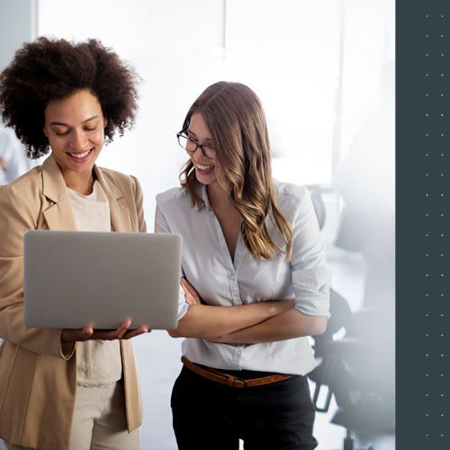 Duas mulheres conversando com o laptop na mão. Estão em um ambiente corporativo.